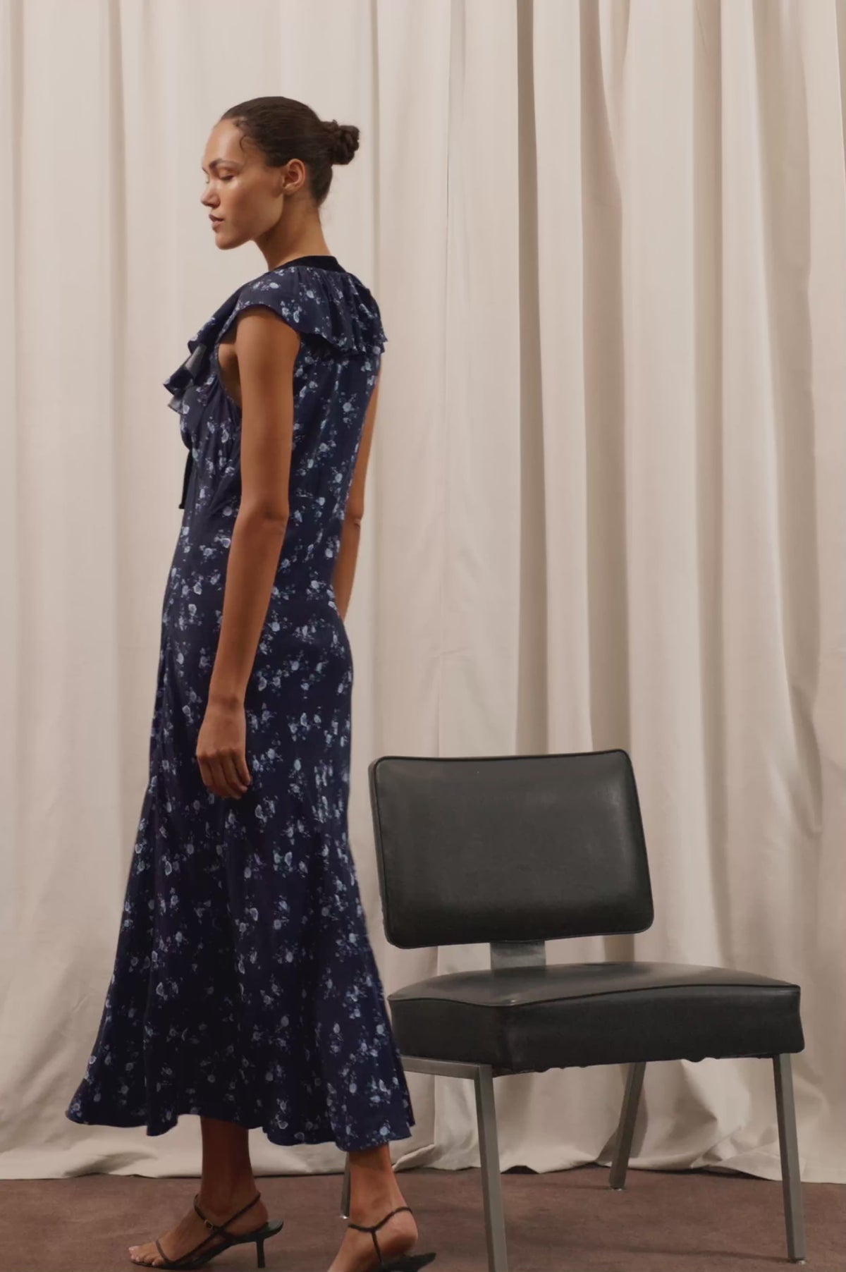 Virginia Maxi Dress in Lightweight Crepe With Ruffle-Trimmed Plunging V-Neckline With Center-Front Bow, Bias Cut, Side Zip Closure, and Godet Insets in Navy Romantic Floral Colorway - Full Body View Featured on Model Posing with Chair (Video).