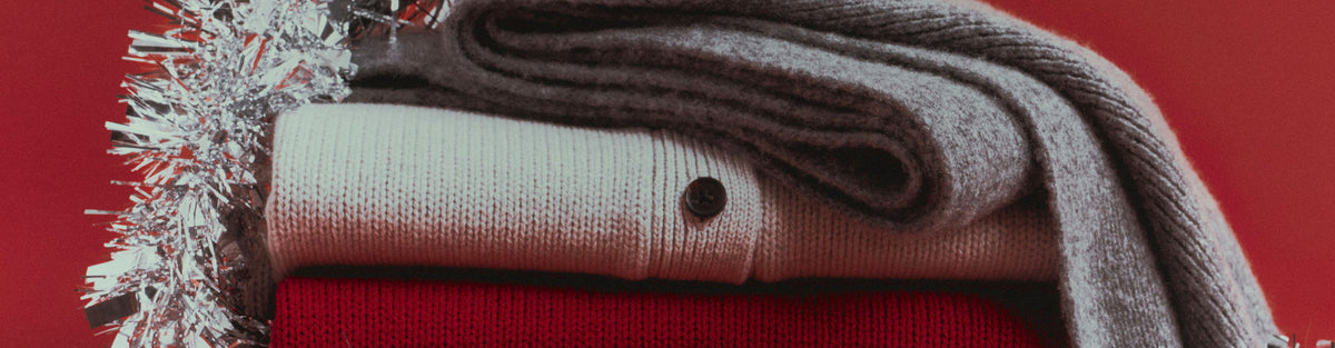A close-up of folded knit textiles in neutral and warm tones, including a gray knit draped over a cream knit, with a red knit base visible beneath and silver tinsel on the left, set against a red background.
