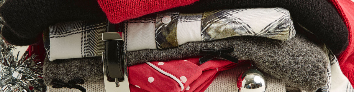 A close-up of folded winter clothing inside a red garment bag, including a plaid shirt, a gray knit sweater, a red top with white polka dots, and a black outer layer, with a silver bell and holiday tinsel visible at the edges.