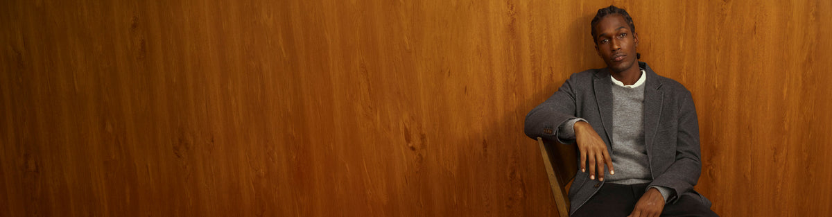 A man wearing a gray blazer over a gray sweater and white shirt sits on a wooden chair against a warm wood-paneled wall.