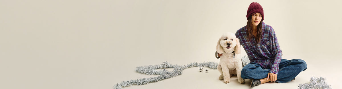 A woman wearing a burgundy knit beanie, purple plaid shirt, and blue jeans sits cross-legged on a light beige studio floor next to a small white dog. Silver tinsel is scattered on the floor around them.
