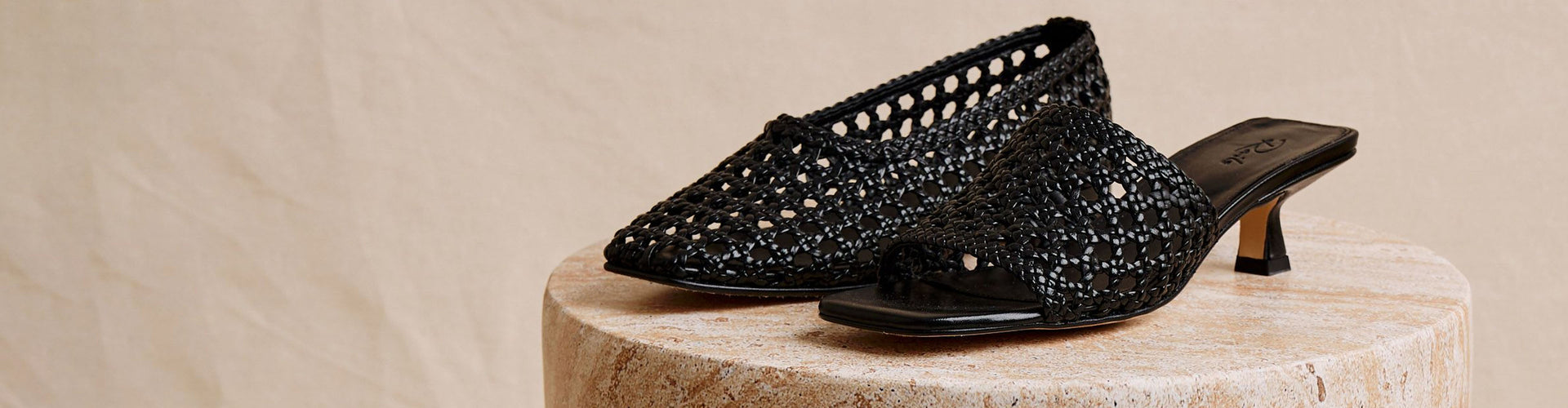A pair of black open-toe mule sandals with a textured, beaded upper and low kitten heels, displayed on a round stone pedestal against a neutral beige background.
