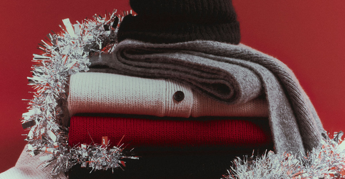 A stack of folded knit winter garments and a dark knit hat wrapped with silver tinsel, set against a solid red background.