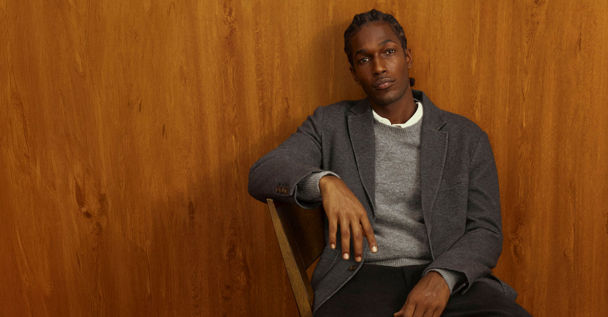 A man wearing a gray blazer over a gray sweater and white collared shirt, sitting against a warm wood-paneled wall.