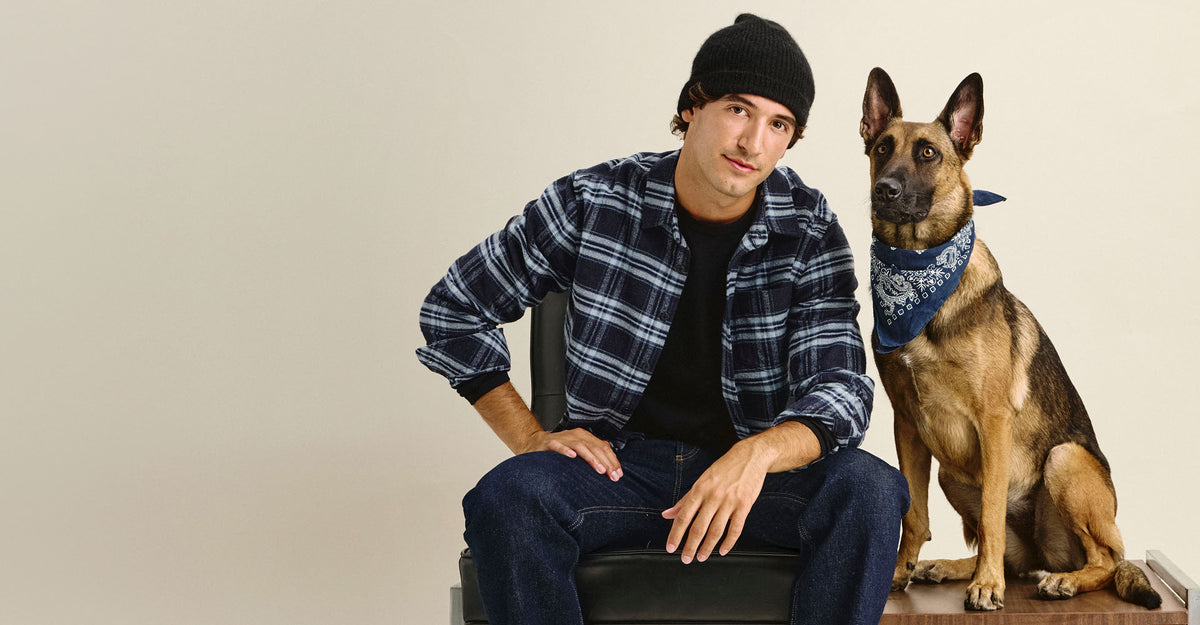 A man wearing a black beanie, a blue and black plaid flannel shirt, and jeans sits on a chair next to a German Shepherd dog wearing a blue bandana, against a plain beige background.