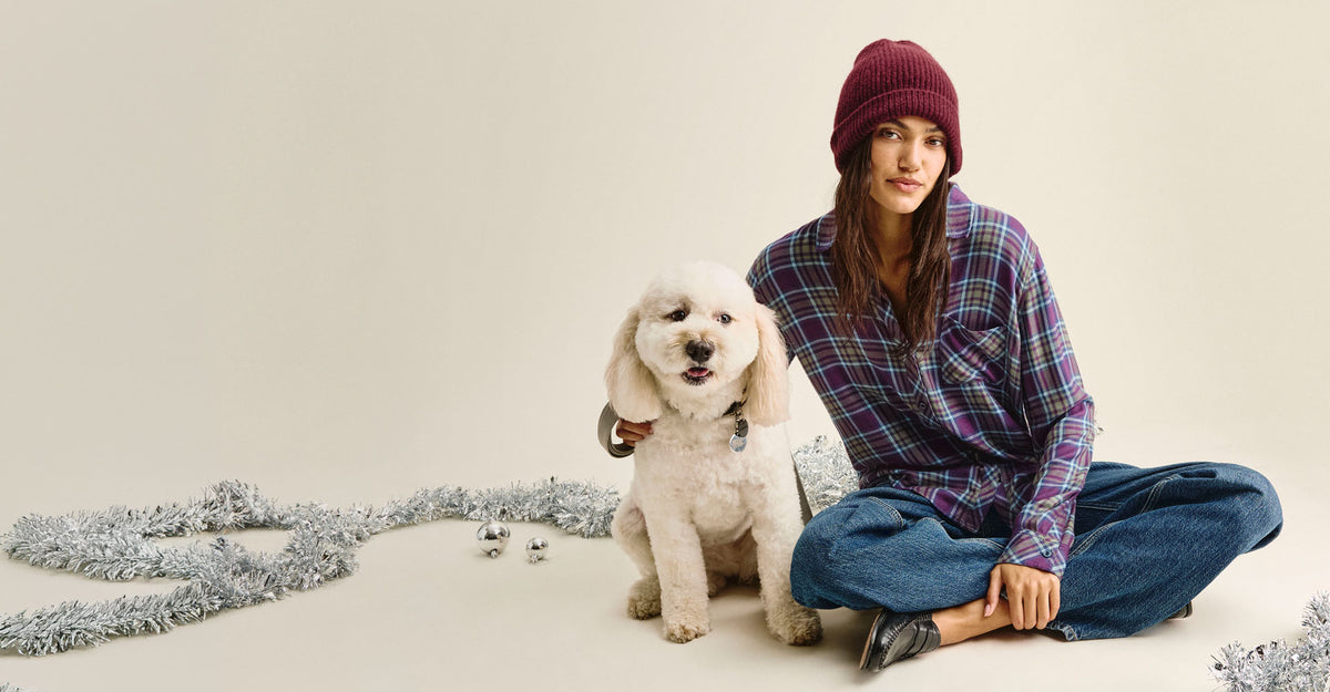A woman wearing a burgundy knit beanie, a purple and blue plaid flannel shirt, and blue jeans sits cross-legged on a cream studio floor next to a small white poodle. Silver tinsel decor is scattered on the floor around them.