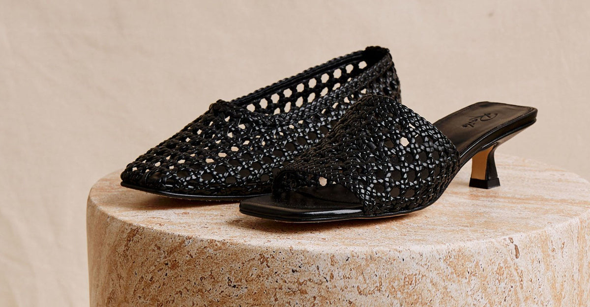 A pair of black open-toe mule sandals with a textured, beaded upper and low kitten heels, displayed on a round stone pedestal against a neutral beige background.