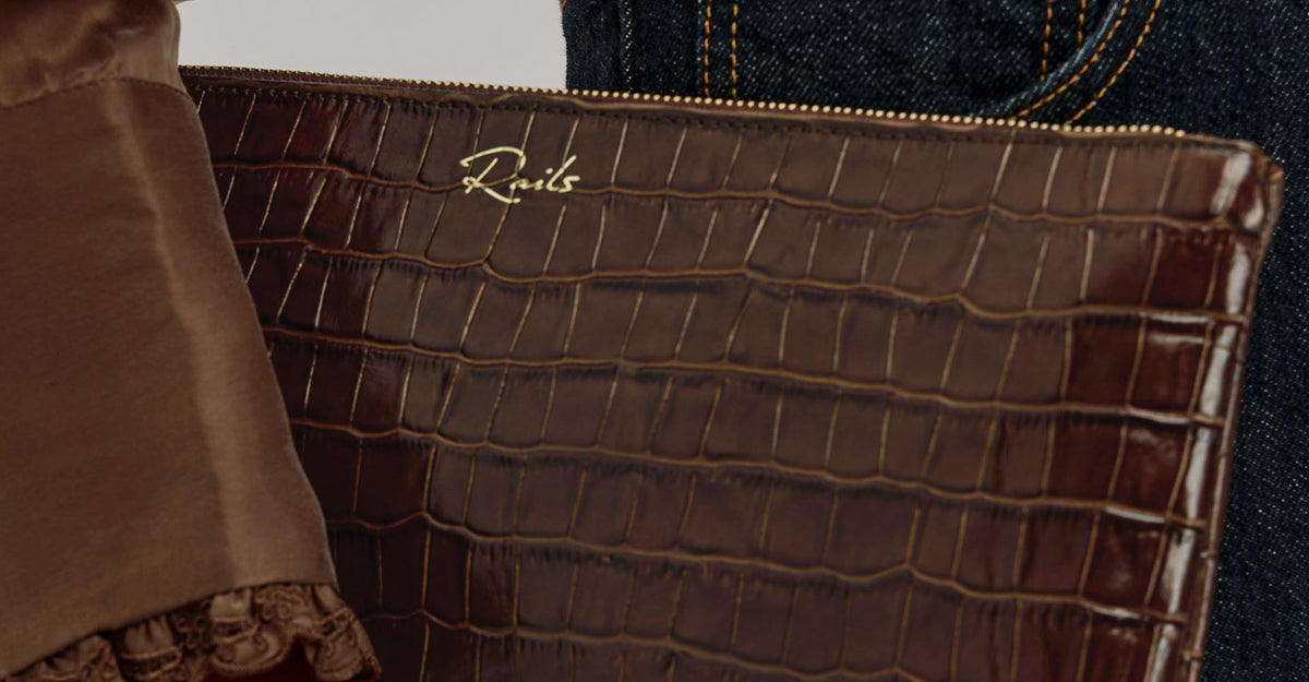 A close-up of a brown crocodile-embossed leather clutch or pouch with a gold script logo reading 'Rav1s' on the front, shown against a neutral background with part of a brown sleeve and dark denim visible.