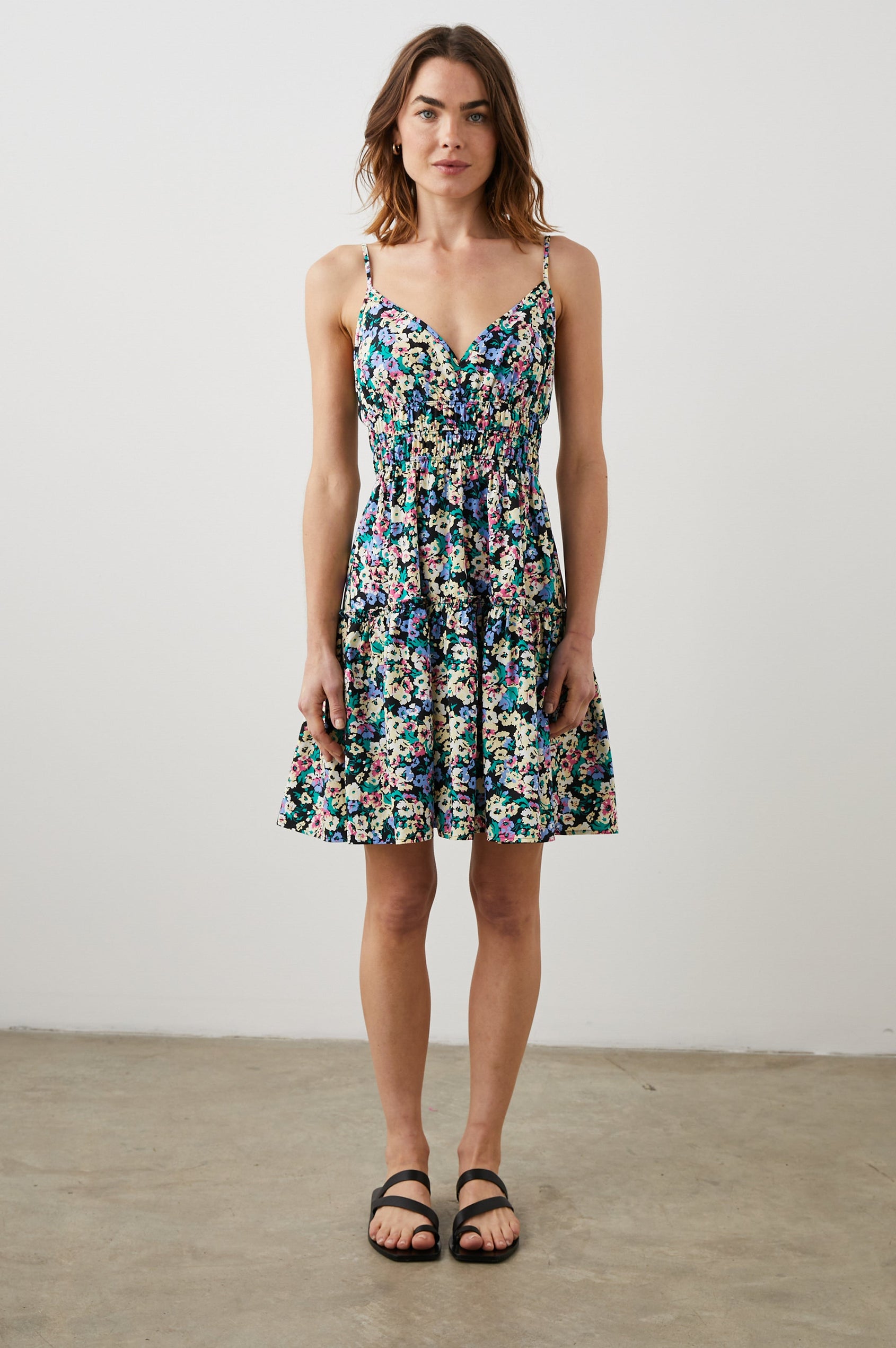 Carmen Sleeveless Mini Dress with Adjustable Spaghetti Straps, a Deep V Neck, a Tiered Skirt and Smocking at the Mid-Bodice in Wildflower Meadow Colorway, Yellow, Purple, Green and Pink on Black - Front Full Body View Featured on Model  