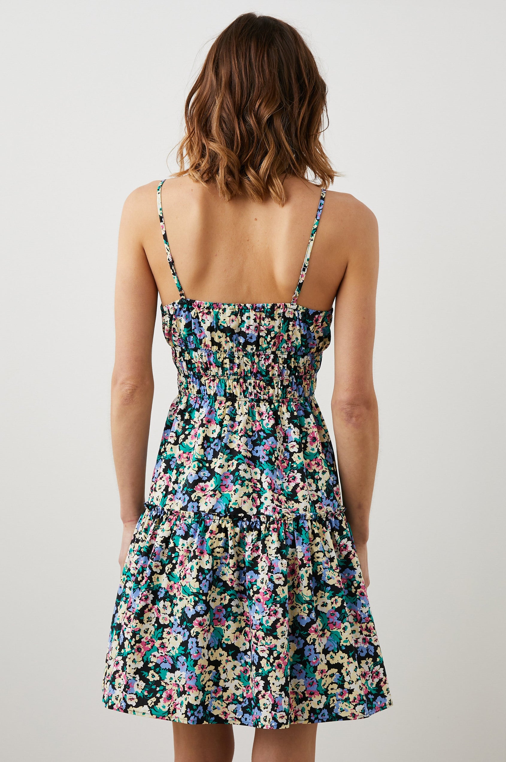 Carmen Sleeveless Mini Dress with Adjustable Spaghetti Straps, a Deep V Neck, a Tiered Skirt and Smocking at the Mid-Bodice in Wildflower Meadow Colorway, Yellow, Purple, Green and Pink on Black - Back View Featured on Model  