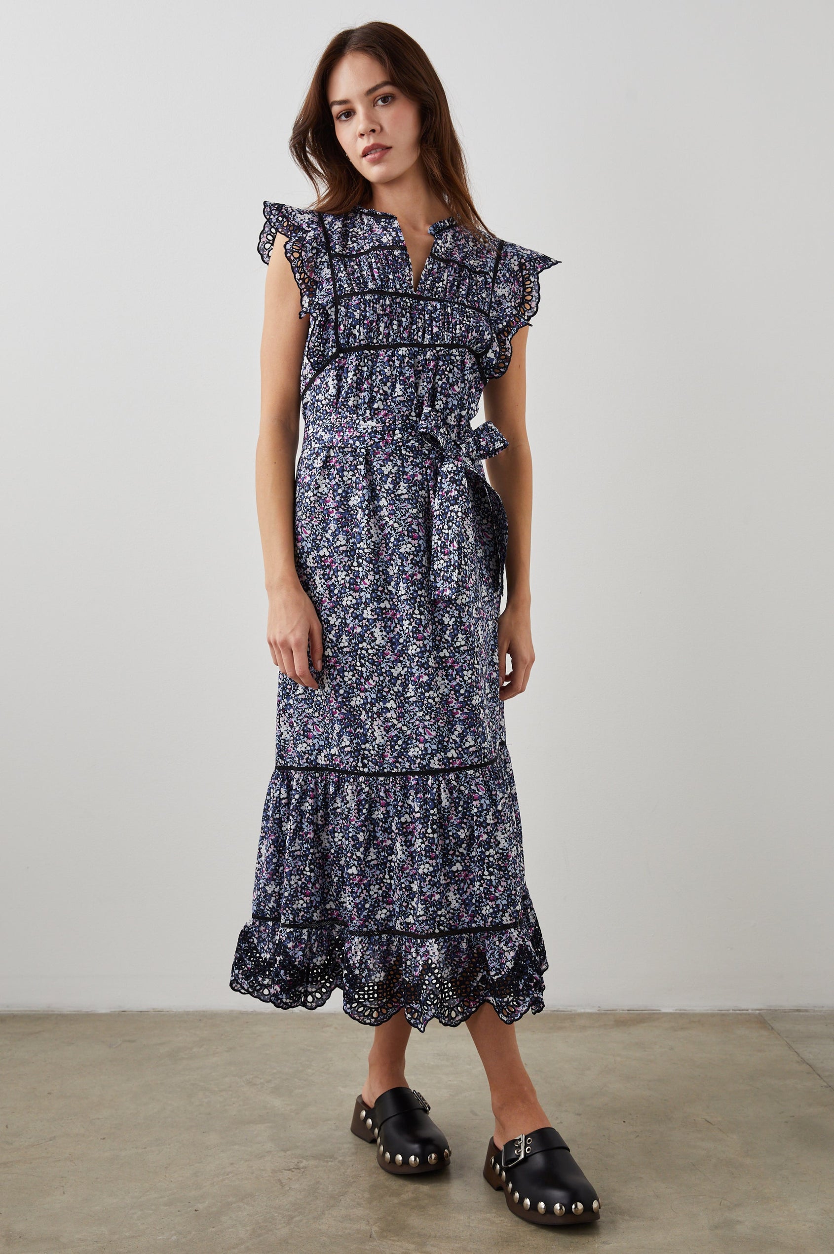 Sofie Short Sleeve Midi Dress with Flutter Sleeves, a V Neck, Panels with Shirring on the Bodice, a Fabric Waist Tie, a Tiered Skirt, and Cutouts on the Sleeves and Along the Hem in Midnight Hyacinth Colorway, Small Flowers in Blues, Pinks and White on Midnight - Front Full Body View Featured on Model 