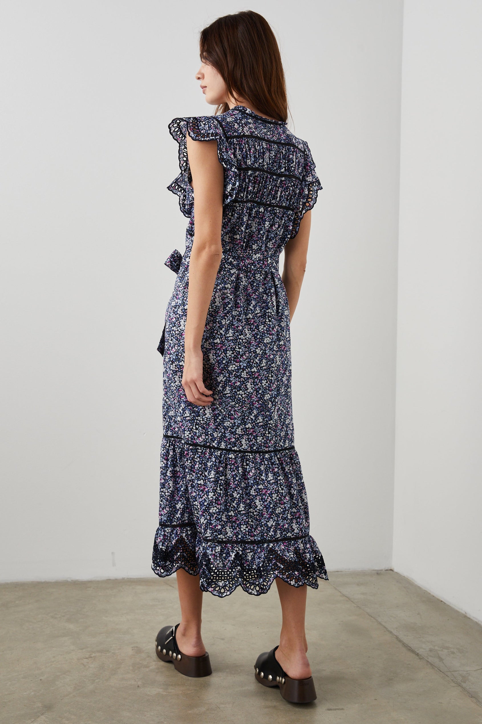Sofie Short Sleeve Midi Dress with Flutter Sleeves, a V Neck, Panels with Shirring on the Bodice, a Fabric Waist Tie, a Tiered Skirt, and Cutouts on the Sleeves and Along the Hem in Midnight Hyacinth Colorway, Small Flowers in Blues, Pinks and White on Midnight - Back Full Body View Featured on Model 